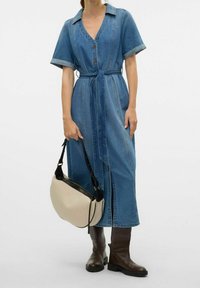 Denimhemdkleid mit kurzen Ärmeln, Knopfleiste, tailliertem Gürtel und seitlichen Schlitzen; kombiniert mit einer hellbeigen Tasche und knielangen braunen Stiefeln.