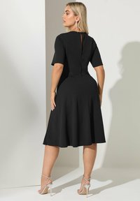 Vestido negro de manga corta con un cuerpo ajustado y falda acampanada. Presenta un detalle de espalda con ojal y una tela suave. Se lleva con sandalias de tiras plateadas.