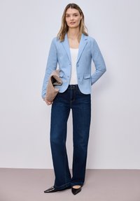 Femme debout contre un mur, portant un blazer bleu clair, un haut blanc, un jean bleu foncé, des chaussures noires, tenant une veste beige pliée dans une main.