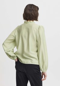 Light green blouse with a textured fabric, featuring a ruffled neckline and puffed sleeves. Worn with black pants, viewed from the back.