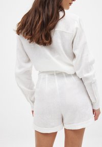 Hvid linnedskjorte med lange ærmer og rygstykke, parret med matchende hvide linnedshorts med opsmøg og selvbinder i taljen.