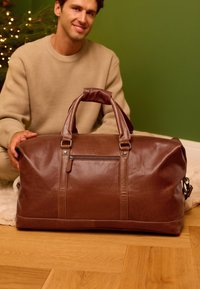 Uomo con maglione beige seduto sul pavimento dietro una grande borsa da viaggio marrone in pelle con tasca con cerniera, muro verde e albero di Natale sfocato sullo sfondo.