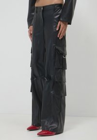 Person, der hochgeschnittene schwarze Leder-Cargo-Hosen mit mehreren Taschen und roten spitzen Absatzschuhen auf einem einfarbigen Hintergrund trägt.