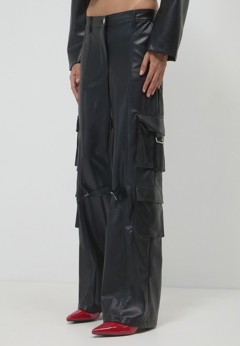 Person, der hochgeschnittene schwarze Leder-Cargo-Hosen mit mehreren Taschen und roten spitzen Absatzschuhen auf einem einfarbigen Hintergrund trägt.