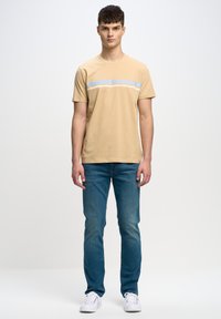 Camiseta de algodón beige de manga corta, con una franja horizontal azul claro y blanca en el pecho, combinada con unos vaqueros azules y unas zapatillas blancas.