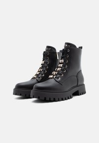 Bottines en cuir noir avec une semelle épaisse, présentant des accents de lacets métalliques et une fermeture éclair sur le côté. Tige texturée avec bout arrondi.