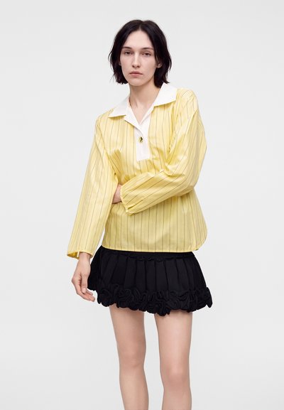Jeune femme aux cheveux courts et foncés portant une chemise jaune à manches longues avec un col blanc et une mini-jupe noire à volants, debout devant un fond uni.