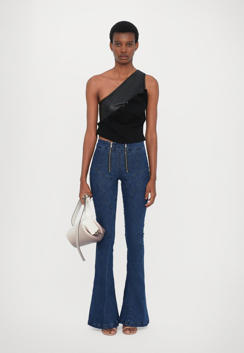 Schwarzes One-Shoulder-Oberteil mit Rüschendetails, kombiniert mit hoch taillierten blauen Jeans mit weitem Bein. Das Model hält eine strukturierte Handtasche in heller Farbe.
