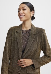 Brauner glitzernder Blazer mit taillierter Passform, Reverskragen und zwei frontalen Taschen, getragen über einem transparenten schwarz-gestreiften Oberteil.