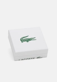 Baltas stačiakampis dėžė su tekstūruota paviršiumi, kurioje yra žalias krokodilo logotipas ir šone spausdinta "LACOSTE" žalia spalva.