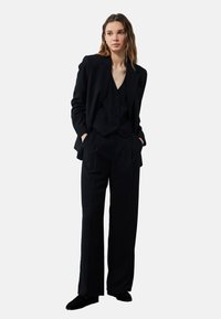 Zwarte op maat gemaakte kostuum bestaande uit een blazer, een vest met knopen en een broek met wijde pijpen, gemaakt van een gestructureerde stof met een gladde afwerking.