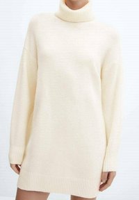 Žena ve volných krémově zbarvených pletených šatech s vysokým límcem, dlouhými rukávy a vroubkovanými manžetami a lemem.