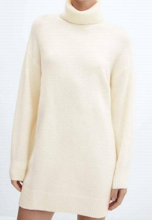 Vrouw in een losse, crème kleur trui jurk met col, lange mouwen en ribbels aan de manchetten en onderrand.