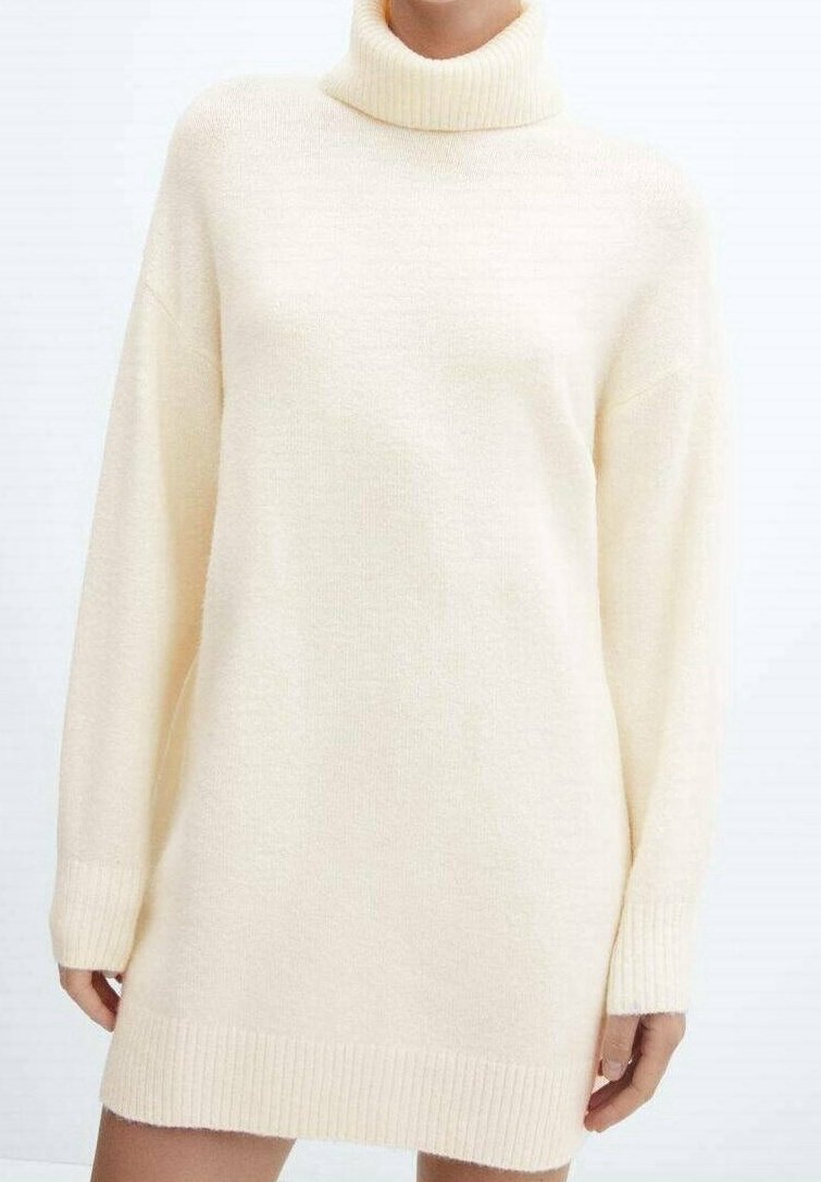 Žena ve volných krémově zbarvených pletených šatech s vysokým límcem, dlouhými rukávy a vroubkovanými manžetami a lemem.