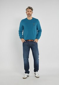 Turkusowy sweatshirt z okrągłym dekoltem i logo na lewej piersi, zestawiony z ciemnoniebieskimi dżinsami oraz białymi sneakersami z czarnymi paskami.