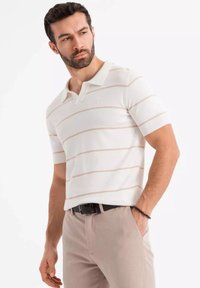 Valkoinen lyhythihainen polo-paita, jossa on vaaleanbeigejä raitoja, neulottu pinta ja resorikaulus. Yhdistetty vaaleanbeigeihin housuihin ja mustaan vyöhön.