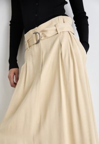 Jupe plissée beige avec une large ceinture et une boucle métallique. Fabriquée dans un tissu lisse, elle tombe élégamment avec un léger éclat.