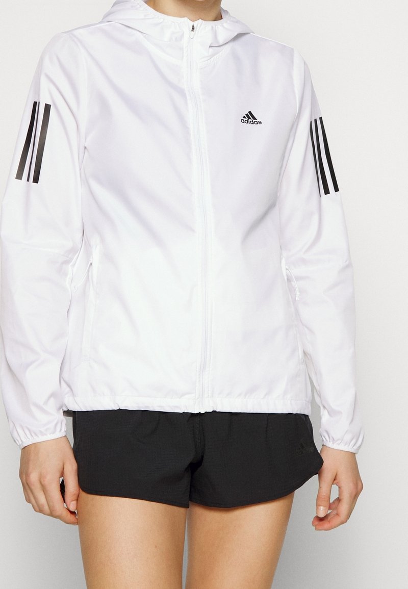 Valkoinen vetoketjullinen takki hupparilla, jossa on mustat kolmi-nauha-yksityiskohdat hihoissa ja Adidas-logo rinnassa. Nauhakuviointi on läsnä.