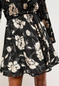 Melna auduma kleita ar ziedu motīvu, kurā redzami smilškrāsas un krēmkrāsas ziedi, papildināta ar sekvīnām un smalku melnu izšuvumu gar malām.