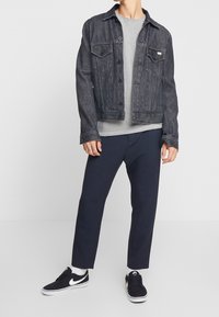 Veste en denim foncé, chemise grise à rayures en dessous, pantalon bleu marine, baskets noires avec des accents blancs. Tenue décontractée aux couleurs contrastées.