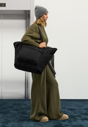 Zwarte gewatteerde totebag met voorvak, gedragen door persoon in olijfgroene outfit en grijze beanie. Glad stof en oversized silhouet.