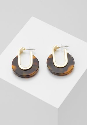 Złote kołowe kolczyki z muszlą żółwia z metalowymi złotymi sztyftami w kształcie litery U na białej, nachylonej powierzchni ekspozycyjnej.