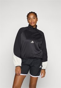 Svart treningsjakke med glidelåskrage og hvite detaljer, sammen med svarte shorts med hvit kant. Materialet er glatt og lett.