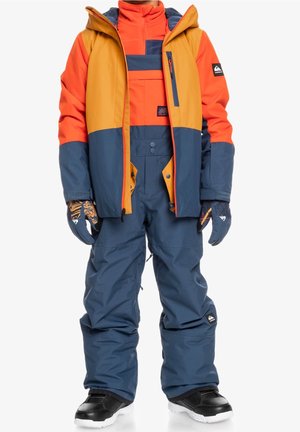 Enfant portant une doudoune d’hiver bicolore orange, jaune et bleue, avec un pantalon de neige bleu assorti, des gants et des après-ski noirs.
