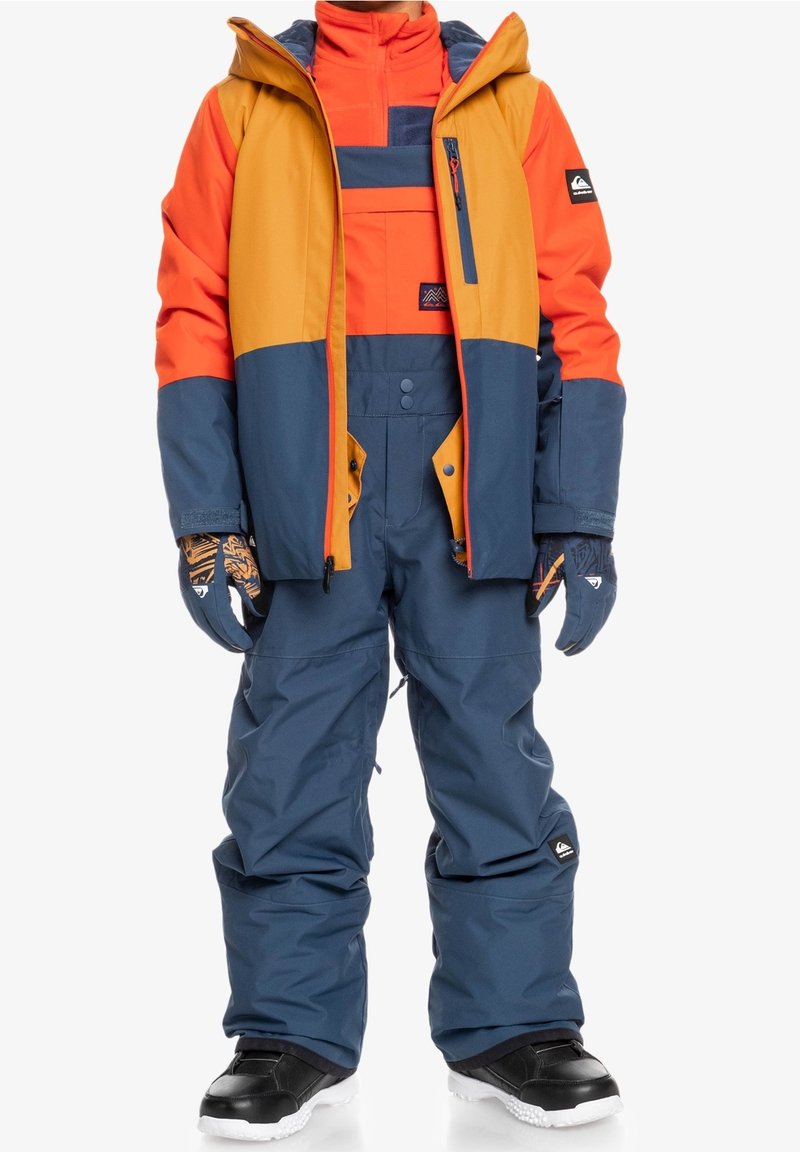 Enfant portant une doudoune d’hiver bicolore orange, jaune et bleue, avec un pantalon de neige bleu assorti, des gants et des après-ski noirs.