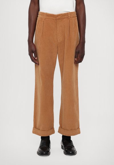 Pantalones de pana marrones con pinzas, pierna ancha y puños en los tobillos. La tela tiene un patrón de ribetes texturizados. Combinados con zapatos negros.