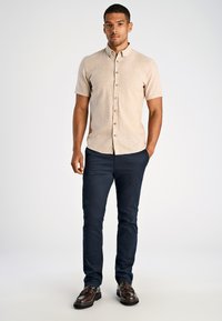 Lichtbeige overhemd met korte mouwen en knopen, gemaakt van geweven stof, gecombineerd met marineblauwe broek en donkerbruine schoenen met een moccasin-stijl accent.