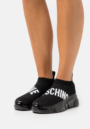 Jambes portant des baskets style chaussettes noires avec des semelles épaisses, inscription blanche « LOVE » sur la chaussure gauche et « MOSCHINO » sur la chaussure droite, sur fond blanc.