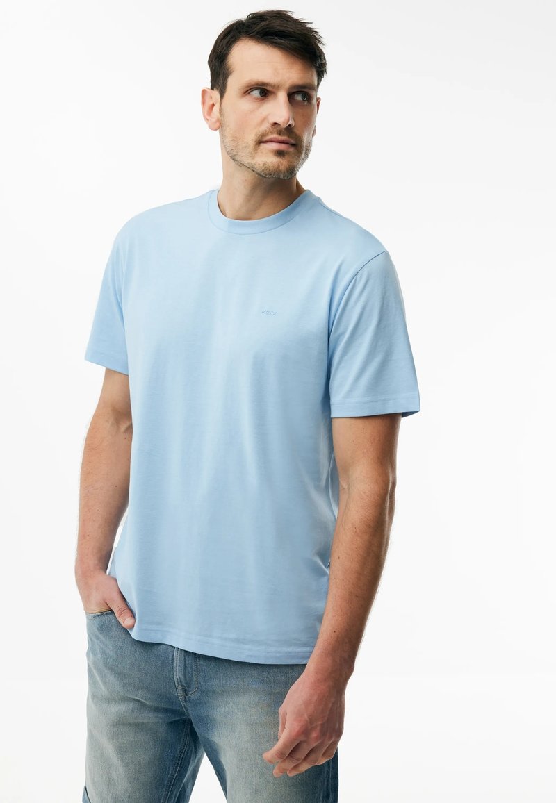 Hombre con camiseta de manga corta azul claro y vaqueros azules descoloridos, de pie con una mano en el bolsillo, mirando hacia un lado sobre fondo blanco.