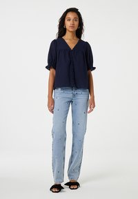 Marineblaue Bluse mit ballongförmigen Ärmeln, Knopfdetails und gewellten Kanten, kombiniert mit hellblauen Jeans, die dekorative Stickereien aufweisen.