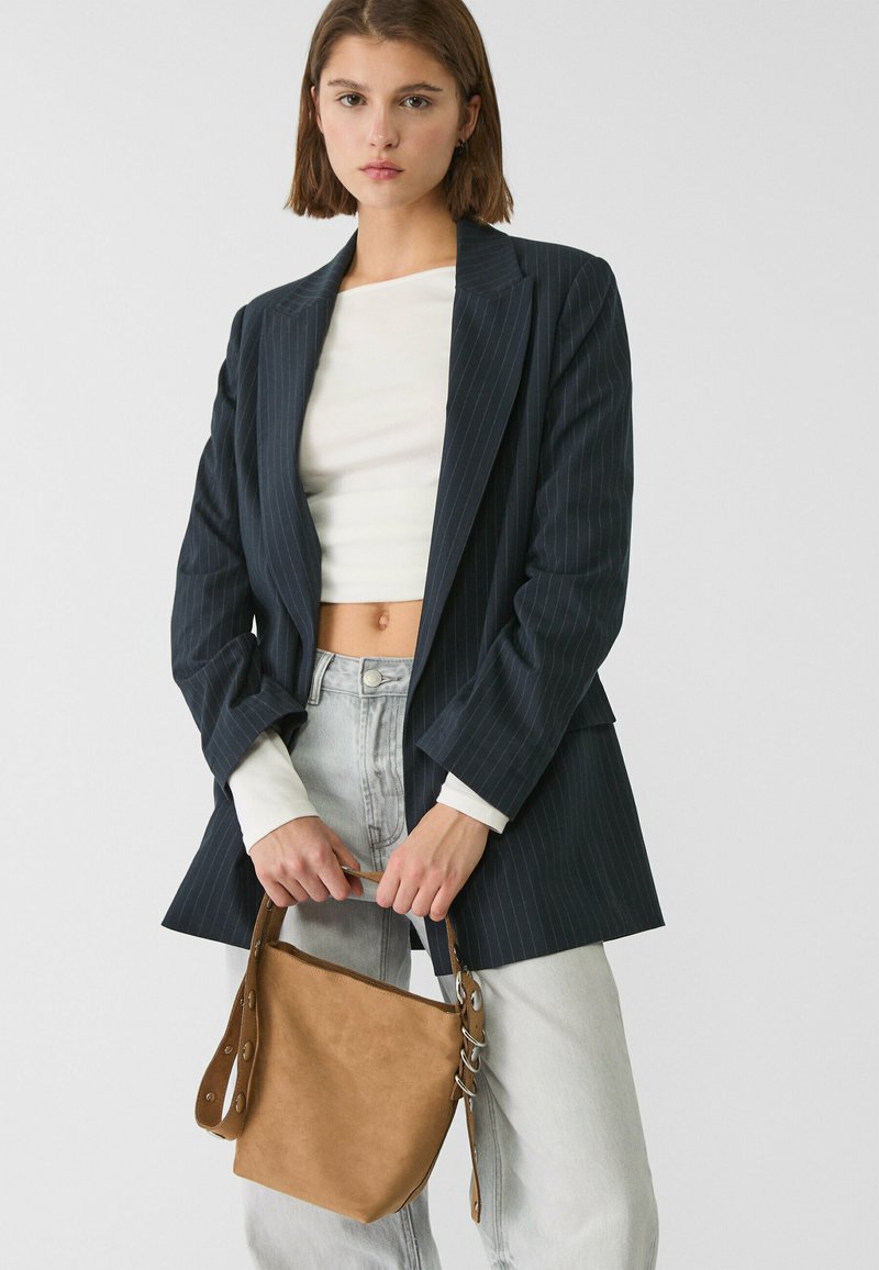 Blazer scuro a righe, crop top bianco, jeans in denim chiaro, e borsa in suede beige con dettagli in argento e tracolla.