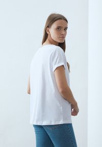 Vit T-shirt med kort ärm och avslappnad passform, med uppvikt ärm och en mjuk textur. Modellen bär den med blå jeans.