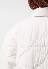 Jachetă albă matlasată cu un design texturat, cu modele curbate, un guler clasic și detalii de cusut vizibile.