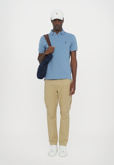 Sininen polo-paita valkoisilla yksityiskohdilla, beige cargo-housut, valkoiset tennarit ja valkoinen lippis, pitäen navy-sinistä laukkua olkapäällä.