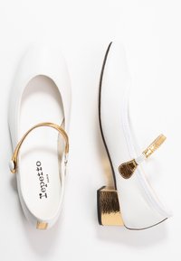 Ballerine bianche con un'accento dorato metallico su un tacco basso, dotate di una fascia e decorazione con bottone, superficie liscia.