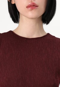 Top bordeaux cu un design texturat, având un guler rotund și un finisaj din material neted, scurtat pentru a expune zona superioară a pieptului.