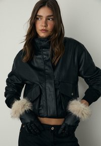 Sort læder cropped jakke med pelsdetaljer på manchetterne, har en høj krave og lynlåslukning. Modellen bærer sorte handsker og mørke jeans.