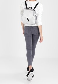 Sac à dos argenté avec fermeture à rabat et détail à cordon de serrage. Confectionné dans un matériau lisse. Porté dans le dos, assorti à un jean gris et des baskets noires.