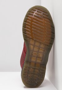 Röd texturerad sko med gummisula, med ett randigt mönster och en logotyp på hälen. Sulan har tydliga greppgropar.