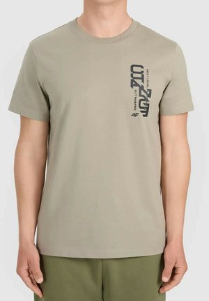 Beżowy t-shirt z krótkim rękawem z czarnym pionowym napisem graficznym na lewej piersi, noszony przez osobę z rękami swobodnie opuszczonymi wzdłuż ciała.