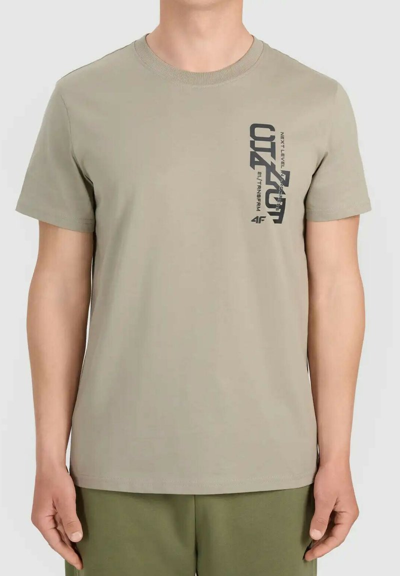 T-shirt beige à manches courtes avec un texte graphique vertical noir sur le côté gauche de la poitrine, porté par une personne avec les bras détendus le long du corps.