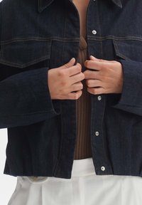 Veste en denim foncé avec une coupe légèrement raccourcie, manches longues, deux poches poitrine et coutures orange contrastantes. Fermeture à boutons sur le devant.