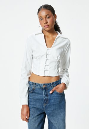 Camicia bianca a maniche lunghe e taglio corto, con chiusura a bottoni frontale, abbinata a jeans blu a vita alta. Tessuto dalla superficie liscia.