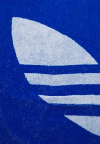 Modrá plážová osuška s textúrovaným povrchom, so stylizovaným bielym logom v tvare veľkého listu a troma horizontálnymi pruhmi.