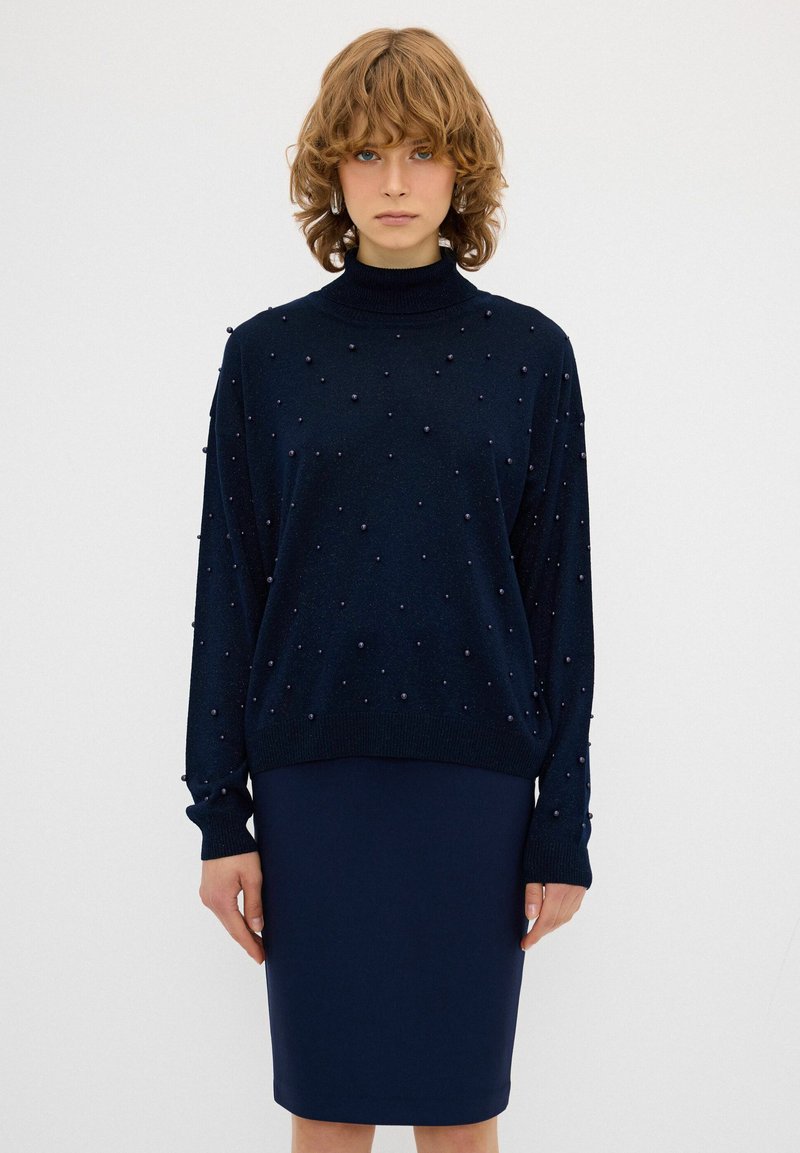 Marineblauer Pullover mit hohem Kragen, verziert mit kleinen dekorativen Perlen. Kombiniert mit einem passenden, figurbetonten marineblauen Bleistiftrock. Weicher Stoff.