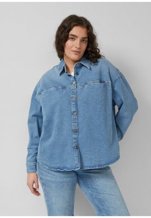 Vrouw met krullend haar, gekleed in een loszittend lichtblauw denim overhemd en bijpassende jeans, tegen een effen grijze achtergrond.
